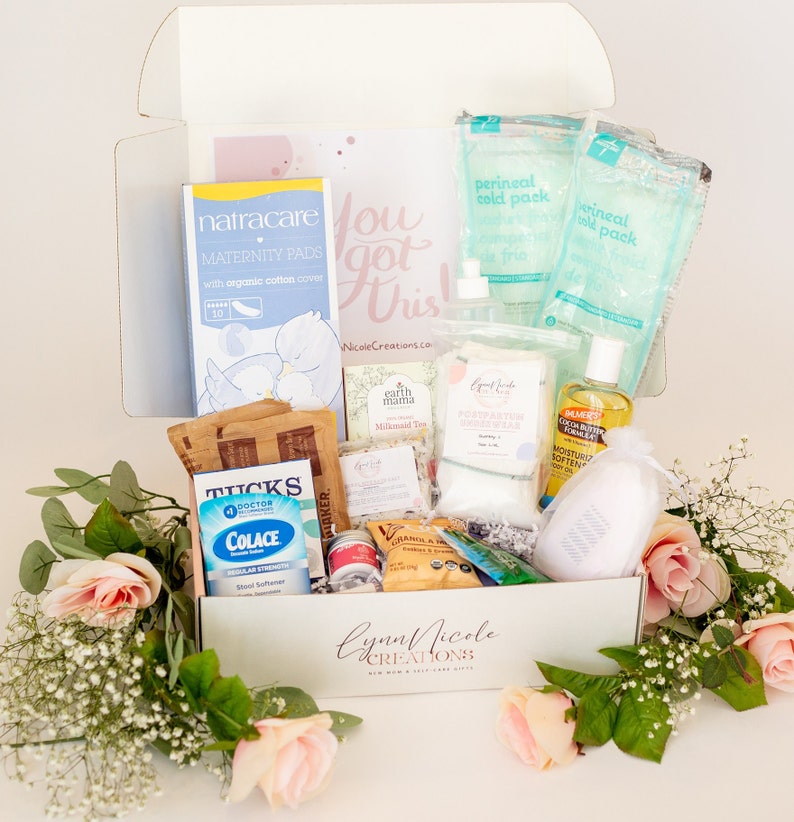 May include: A white gift box filled with various postpartum care items, including perineal cold packs, maternity pads, a stool softener, and a bottle of cocoa butter. The box is decorated with pink roses and the text "Lynn Nicole Creations New Mom & Self Care Gifts".