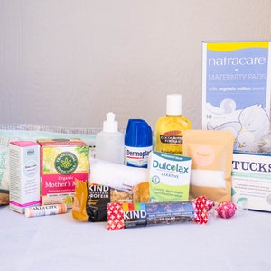 May include: A collection of postpartum recovery items, including pads, wipes, tea, snacks, and a perineal cold pack. The items are arranged on a white surface.