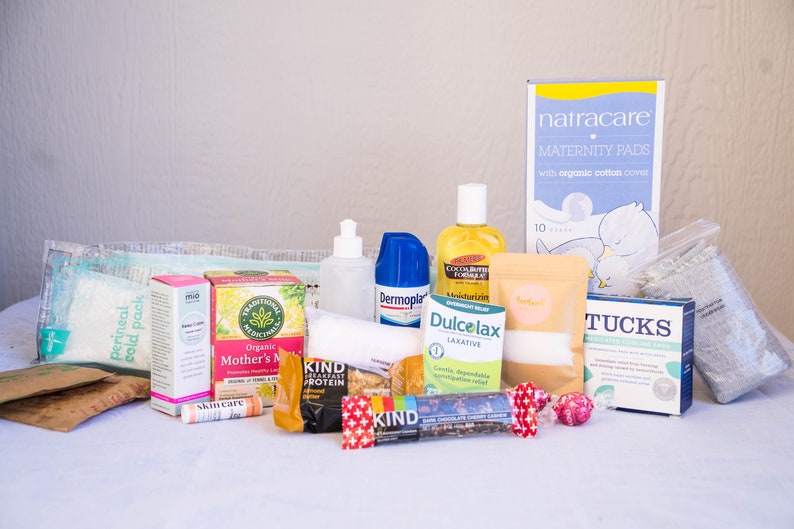 May include: A collection of postpartum recovery items, including pads, wipes, tea, snacks, and a perineal cold pack. The items are arranged on a white surface.