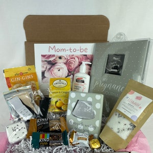 May include: A gift box filled with items for a new mom-to-be. The box contains a pregnancy journal, bath salts, tea, snacks, lotion, and a framed ultrasound photo. The box is decorated with a pink ribbon and the words "Mom-to-be".