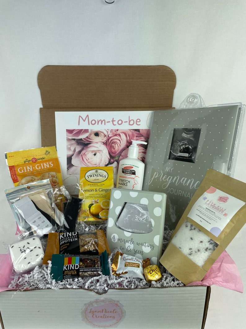 May include: A gift box filled with items for a new mom-to-be. The box contains a pregnancy journal, bath salts, tea, snacks, lotion, and a framed ultrasound photo. The box is decorated with a pink ribbon and the words "Mom-to-be".