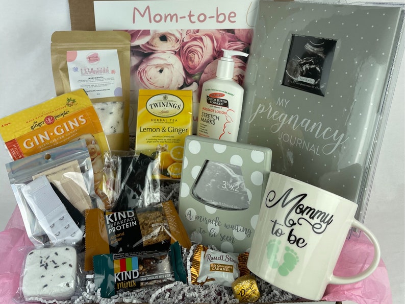 May include: A gift box filled with items for a new mom, including a pregnancy journal, a mug that says "Mommy to be", tea, bath products, and snacks.