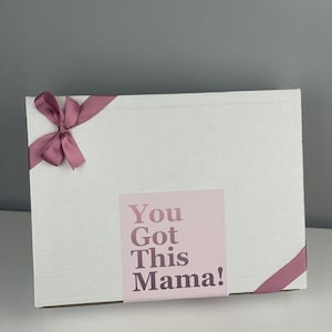 May include: A rectangular white cardboard box decorated with a dusty rose ribbon tied in a bow on the top left corner. A square label is centred on the front, featuring the text "You Got This Mama!" in a dark purple font on a light pink background.