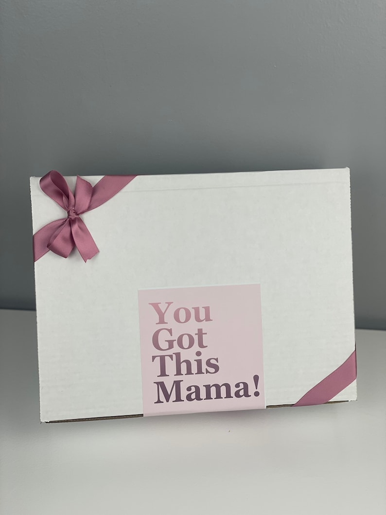 May include: A rectangular white cardboard box decorated with a dusty rose ribbon tied in a bow on the top left corner. A square label is centred on the front, featuring the text "You Got This Mama!" in a dark purple font on a light pink background.
