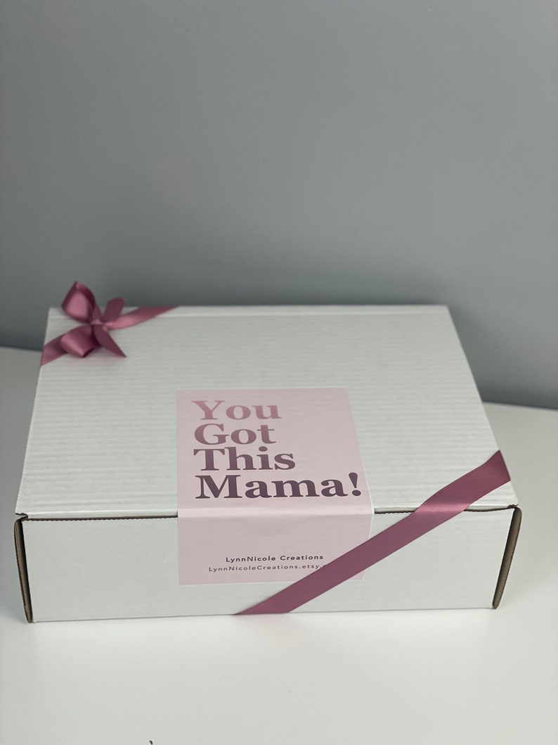 May include: A white rectangular cardboard shipping box wrapped with a mauve satin ribbon tied in a bow. A rectangular label on the lid features the text "You Got This Mama!" in dark purple, with "LynnNicole Creations" printed below.