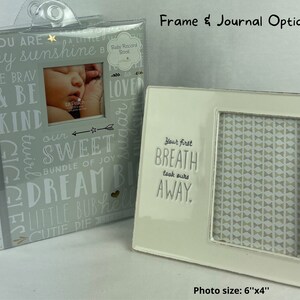 May include: A white ceramic frame with a gray and white patterned background. The frame has a cutout for a 6x4 inch photo and the text "Your first breath took ours away." The frame is next to a gray journal with the text "You are a little sunshine" and other positive words.