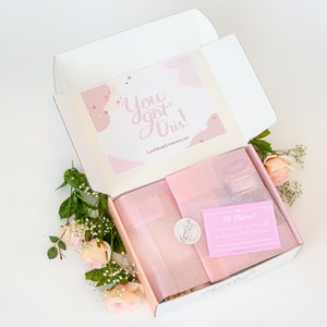 May include: An open white box with pink tissue paper and a pink card that says "Hi There!" inside. The box is decorated with a pink and white card that says "You got this!" and has a pink and white abstract design. The box is surrounded by pink roses and green leaves.