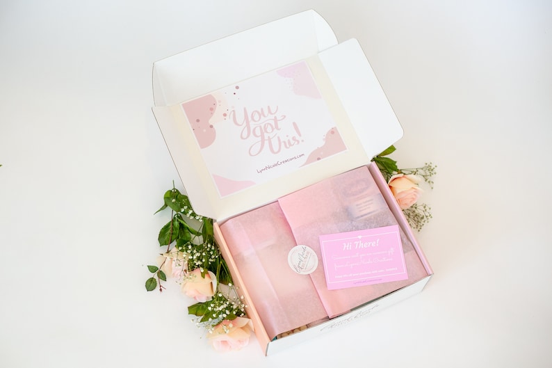 May include: An open white box with pink tissue paper and a pink card that says "Hi There!" inside. The box is decorated with a pink and white card that says "You got this!" and has a pink and white abstract design. The box is surrounded by pink roses and green leaves.