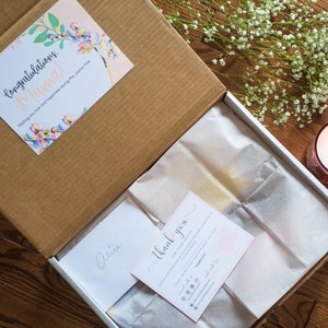 May include: A cardboard gift box containing white tissue paper, a card with the text Congratulations Mama, a thank you note, and a small envelope labelled Alice. The box is decorated with floral accents and accompanied by a lit candle and baby's breath.