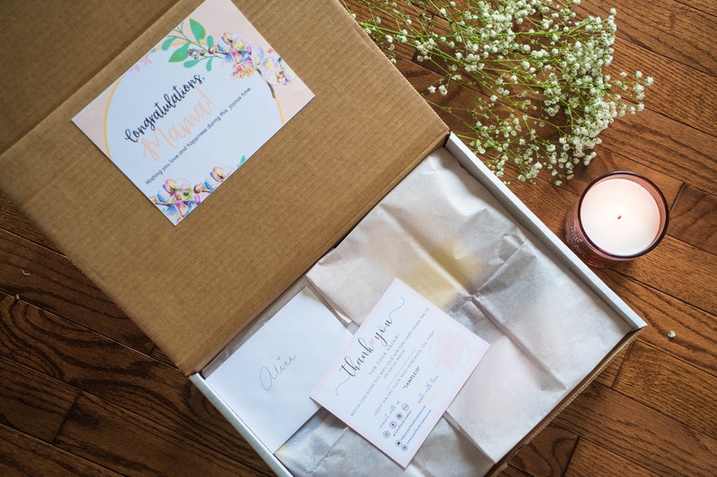 May include: A cardboard gift box containing white tissue paper, a card with the text Congratulations Mama, a thank you note, and a small envelope labelled Alice. The box is decorated with floral accents and accompanied by a lit candle and baby's breath.
