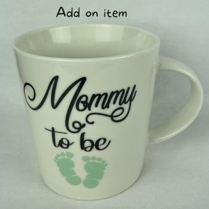 May include: White ceramic mug with black text that reads "Mommy to be" and two green baby footprints.