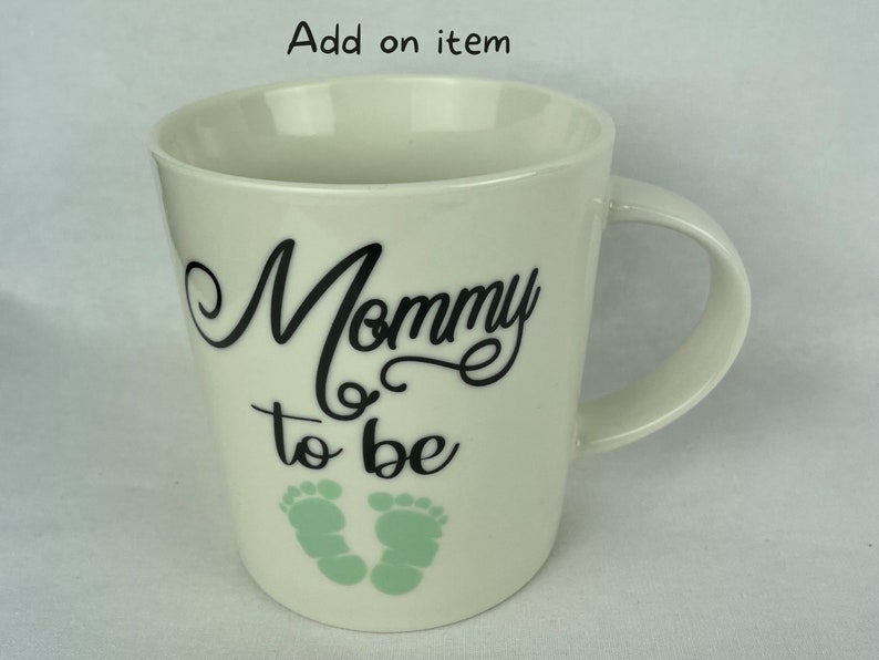 May include: White ceramic mug with black text that reads "Mommy to be" and two green baby footprints.