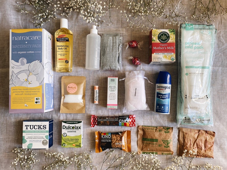 May include: A flat lay of various items for a postpartum recovery kit, including maternity pads, body oil, a cold pack, tea, and snacks.