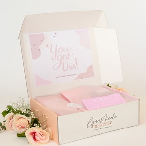 May include: A pink and white box with the text "You got this!" on the lid. The box is open and contains a pink and white gift bag and a pink card with the text "The Journey". The box is labeled "Lynn Nicole Creations" and has a pink and white floral design.