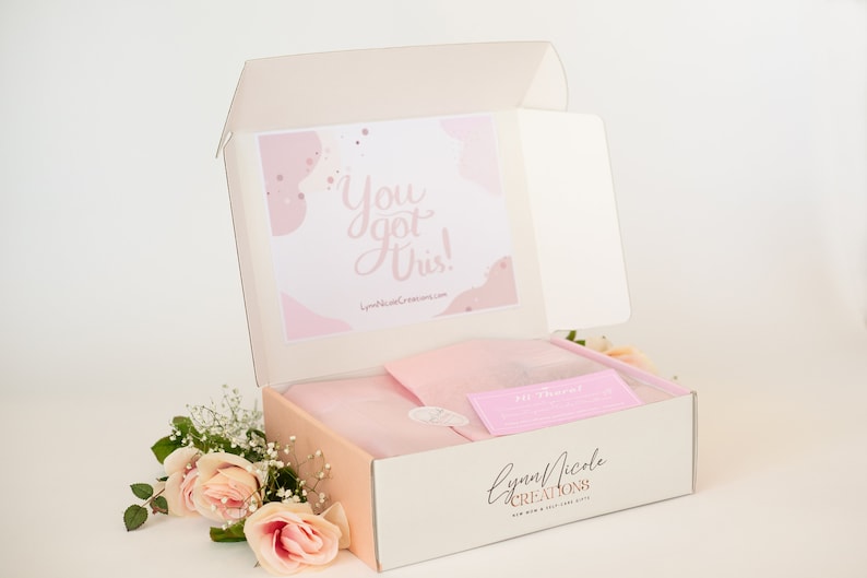 May include: A pink and white box with the text "You got this!" on the lid. The box is open and contains a pink and white gift bag and a pink card with the text "The Journey". The box is labeled "Lynn Nicole Creations" and has a pink and white floral design.