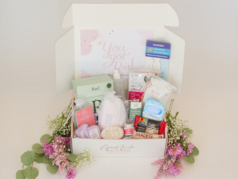 May include: A white box filled with various self-care items, including bath bombs, body scrub, and skincare products. The box is decorated with pink flowers and the text "You got this!"  The box is labeled "Lynette Nicole Creations".
