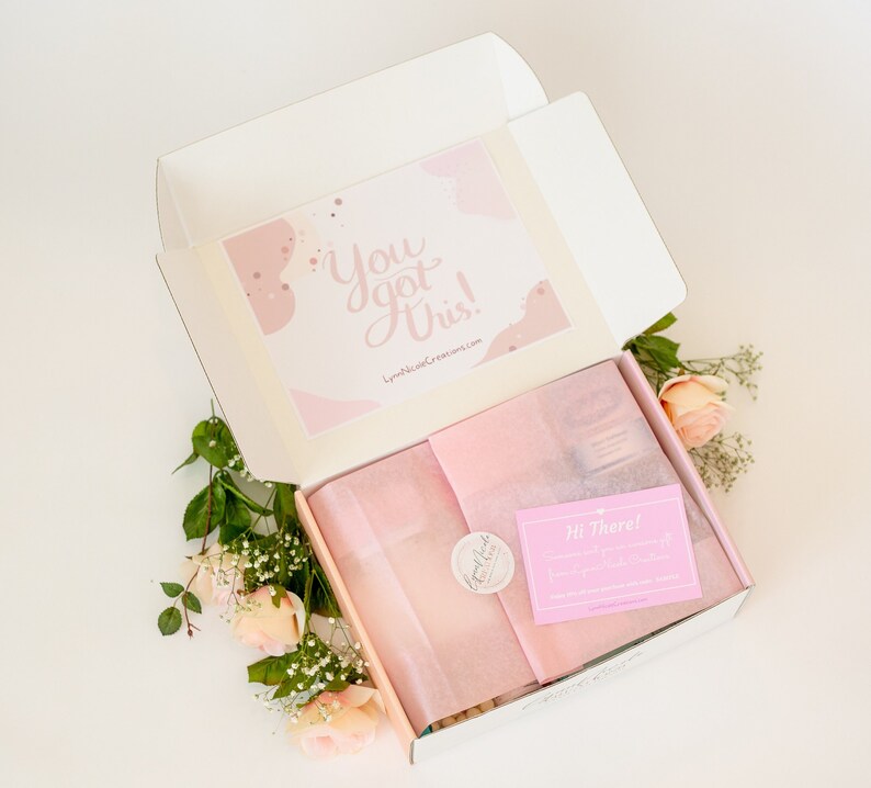 May include: An open white box with pink tissue paper and a pink card that says "You got this!"  The box is surrounded by pink roses and greenery.