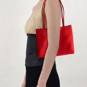 May include: A rectangular shoulder bag crafted from a vibrant red satin fabric. The bag features a smooth, lustrous finish and two thin, matching red shoulder straps. The design is simple and structured, with a clean, minimalist rectangular shape.
