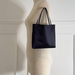 May include: A small, black tote bag with two thin handles. The bag is rectangular and appears to be made of a smooth, possibly satin-like material. The bag is displayed on a mannequin.