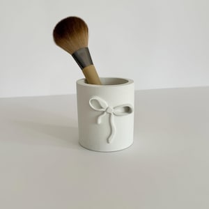 May include: White ceramic makeup brush holder with a raised bow design. A brown makeup brush is in the holder.