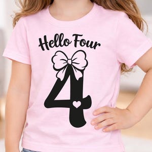 Könnte beinhalten: Hellrosa T-Shirt mit dem schwarzen Schriftzug "Hello Four". Darunter befindet sich eine große schwarze Zahl 4, verziert mit einer weißen Schleife und einem kleinen Herz. Das T-Shirt hat kurze Ärmel.