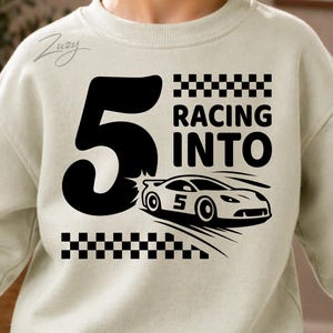 May include: Cream-colored sweatshirt with a black graphic. The graphic features a large number 5, the words "RACING INTO", a race car, and a checkered flag design. The sweatshirt is likely for a child's birthday.
