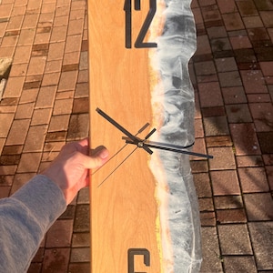 May include: A rectangular wooden wall clock with black numerals at the 12 and 6 positions. The right edge features a resin design with white, grey, and gold tones. Black clock hands indicate the time.