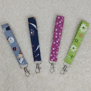 Könnte beinhalten: Vier Stoffschlüsselanhänger mit verschiedenen Mustern und silbernen Schlüsselringen. Die Schlüsselanhänger sind blau mit Sternen, rosa mit Blumen, grün mit Blumen und grau mit einem weißen Tiermuster.