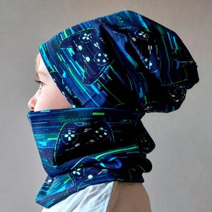 Könnte beinhalten: Ein dunkelblaues Beanie und ein passender Schlauchschal mit einem wiederkehrenden Muster aus schwarzen Videospiel-Controllern, akzentuiert durch digitale Glitch-Linien und Lichtstreifen in Cyan und Limettengrün auf dem Stoff.