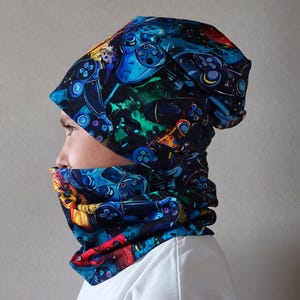 Könnte beinhalten: Eine Mütze und ein passender Schlauchschal mit einem lebendigen, mehrfarbigen Grafikdruck von Videospiel-Controllern auf dunklem Hintergrund mit spritzartigen Texturen in Blau, Grün, Rot und Gelb auf der gesamten Stoffoberfläche.