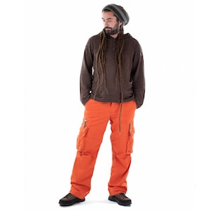 Pode incluir: Um homem vestindo um suéter marrom com capuz, dreadlocks longos, um gorro cinza e calças cargo laranja.