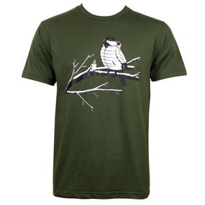 May include: Olive green t-shirt featuring a graphic of a bird in a hooded jacket sitting on a tree branch. A small white coffee cup sits next to the bird. The bird and branch are in black and white.