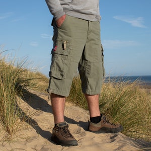 May include: A man wearing green cargo shorts and brown hiking boots stands on a sand dune. The shorts have a large cargo pocket on the left leg and a smaller pocket on the right leg.