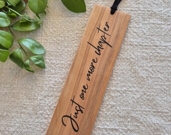May include: A wooden bookmark with the words "Just one more chapter" in black script. It has a black cord and a natural wood grain. The bookmark is on a textured, light-coloured surface. The bookmark is rectangular in shape.