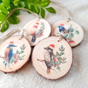 May include: Four wooden ornaments featuring different Australian birds wearing Santa hats. Each ornament is round and has a natural wood grain finish. The birds are painted in realistic detail and are surrounded by green leaves and red berries.