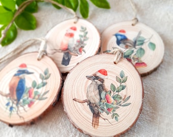 May include: Four wooden ornaments featuring different Australian birds wearing Santa hats. Each ornament is round and has a natural wood grain finish. The birds are painted in realistic detail and are surrounded by green leaves and red berries.