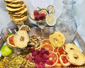Pode incluir: Uma bandeja dourada com uma variedade de fatias de frutas secas, incluindo abacaxi, laranja e kiwi. Também há framboesas e uma jarra de vidro transparente com uma rolha.