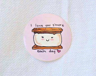 May include: A round, light pink button with a hand-drawn illustration of a s'more. The s'more has a smiling face and the text "I love you s'more each day" in black lettering. A small pink heart is at the end of the text.