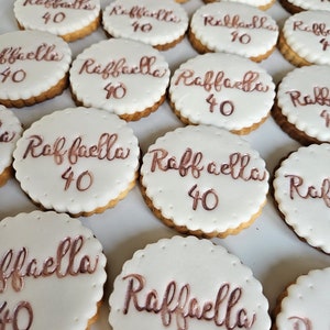 Puede incluir: Una colección de galletas redondas con glaseado blanco y el nombre "Raffaella" y el número "40" escritos en una escritura color cobre. Las galletas tienen un borde festoneado y una base dorada.