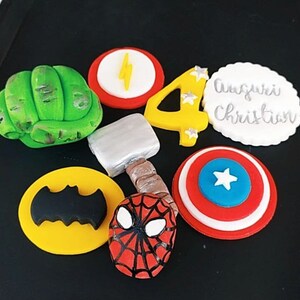 Può includere: Un set di topper per torte a tema supereroi, tra cui un pugno verde di Hulk, un fulmine giallo su un cerchio rosso, un numero quattro giallo con stelle, un cerchio bianco con il testo "Auguri Christian", un martello argentato, uno scudo di Capitan America rosso e blu, un cerchio giallo con un simbolo di Batman nero e una maschera di Spider-Man rossa e nera.
