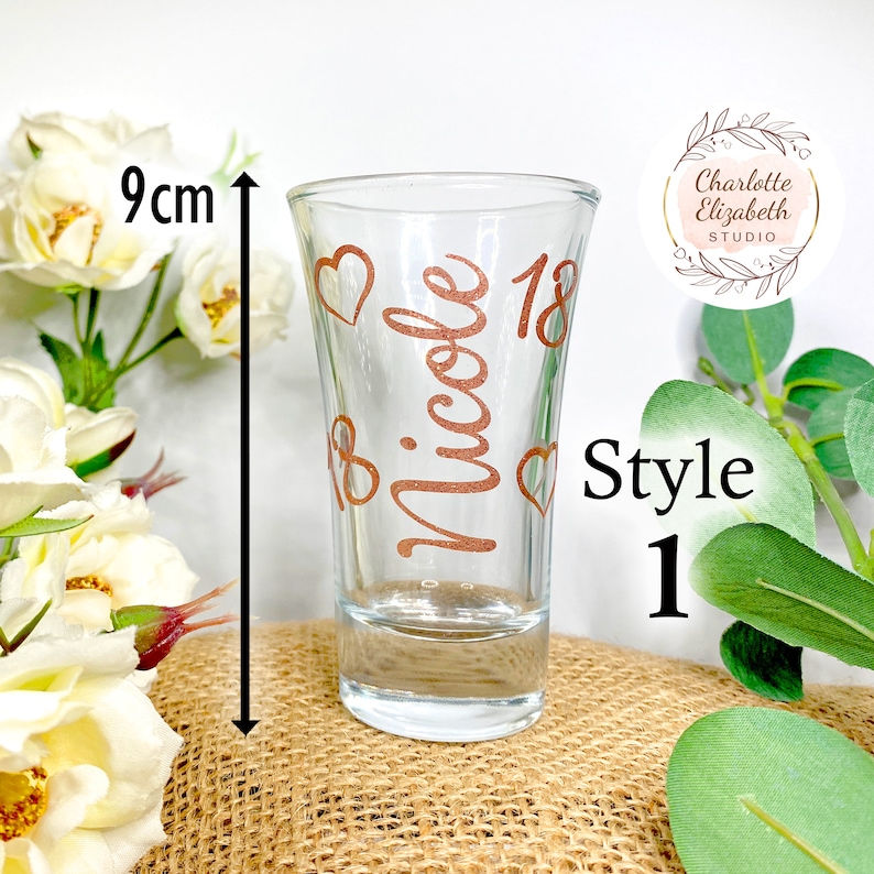 May include: A clear glass shot glass with a copper-colored design that says "Nicole 18" with hearts. The shot glass is 9 cm tall. Style 1.