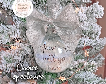 May include: A clear, spherical glass Christmas ornament filled with white faux snow, featuring a large, shimmering silver fabric bow at the top. The ornament displays the black text "Your text will go here" on its front surface.