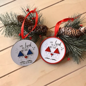 Op de afbeelding: Twee ronde houten ornamenten met een rode en witte rand. Elk ornament heeft een rood hart, twee huizen en de tekst "Ik wens dat je naast de deur woonde". Een ornament heeft een blauw huis en de tekst "zodat ik je vaker kon zien".