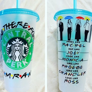 May include: A white reusable cup with a green Starbucks logo and the words "Central Perk" and "There for you" written on it. The cup also has the words "I'm" and "Friends" written on it with colorful dots. The cup has a blue lid and a blue straw.