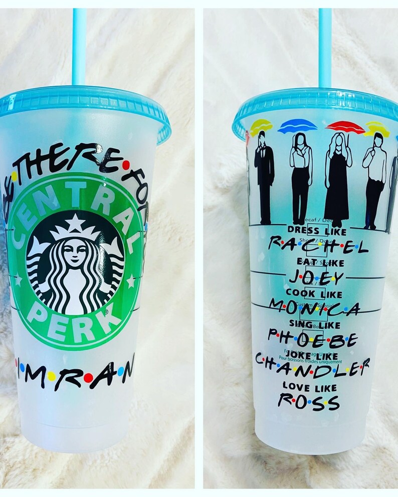 May include: A white reusable cup with a green Starbucks logo and the words "Central Perk" and "There for you" written on it. The cup also has the words "I'm" and "Friends" written on it with colorful dots. The cup has a blue lid and a blue straw.