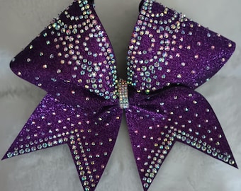 May include: Purple cheer bow with iridescent rhinestones. The bow is made of glitter fabric and has a pointed shape.