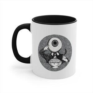May include: A white ceramic coffee mug with a black rim and handle. The mug features a black and white illustration of an eye with a body and a spoon, eating a bowl of brains. The illustration is surrounded by a circle with a textured background.