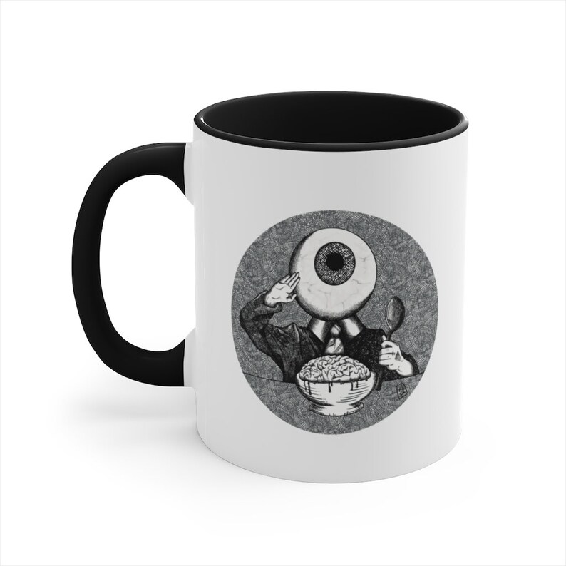 May include: A white ceramic coffee mug with a black rim and handle. The mug features a black and white illustration of an eye with a body and a spoon, eating a bowl of brains. The illustration is surrounded by a circle with a textured background.
