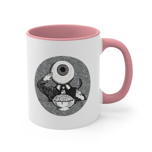 May include: White ceramic coffee mug with a pink rim. The mug features a black and white illustration of a person with an eye for a head eating cereal.