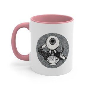 May include: White ceramic coffee mug with a pink rim. The mug features a black and white illustration of an eye with a body, wearing a suit and saluting, eating a bowl of brains.